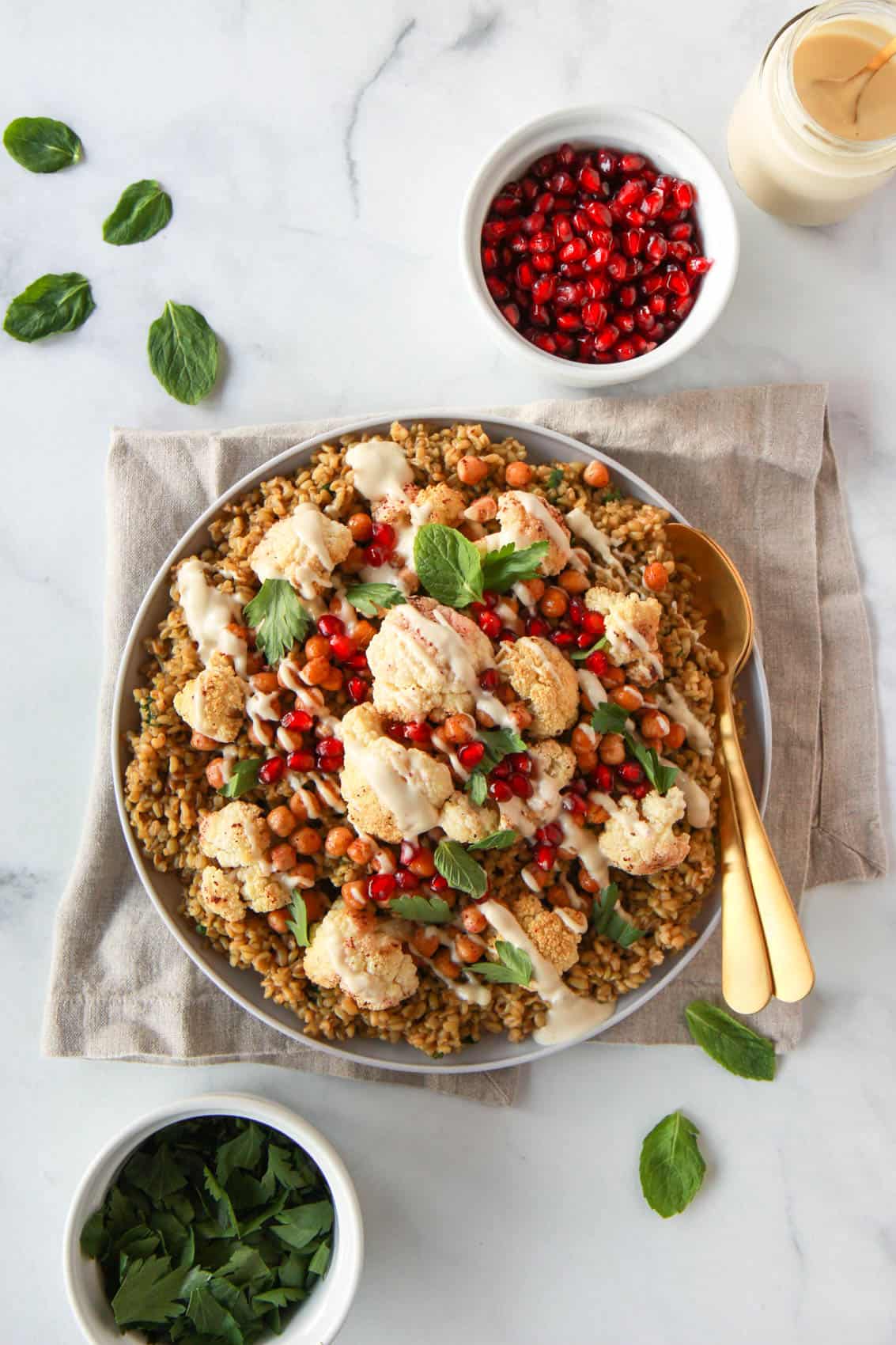 This image shows an overhead shot of the freekeh dish being served alongside fresh parsley, pomegranate seeds, and tahini sauce.