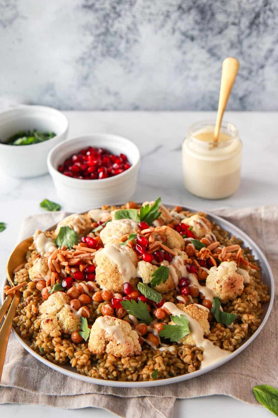 The final dish is plated, with roasted cauliflower and chickpeas over the cooked freekeh, showing the tahini in the background which is drizzled over the dish.