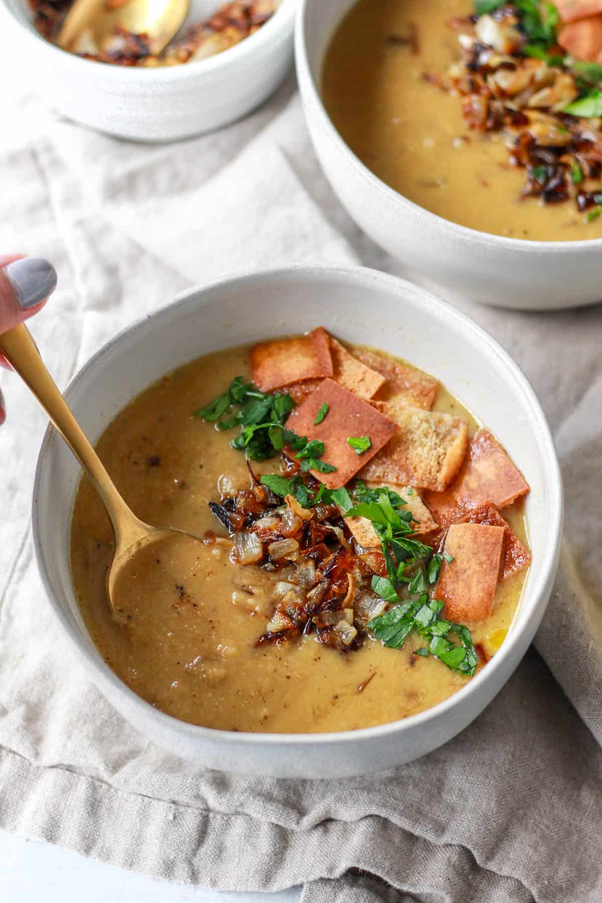 A spoon dipping into a bowl of Arabic lentil soup topped with pita chips, crispy onions, and parsley.