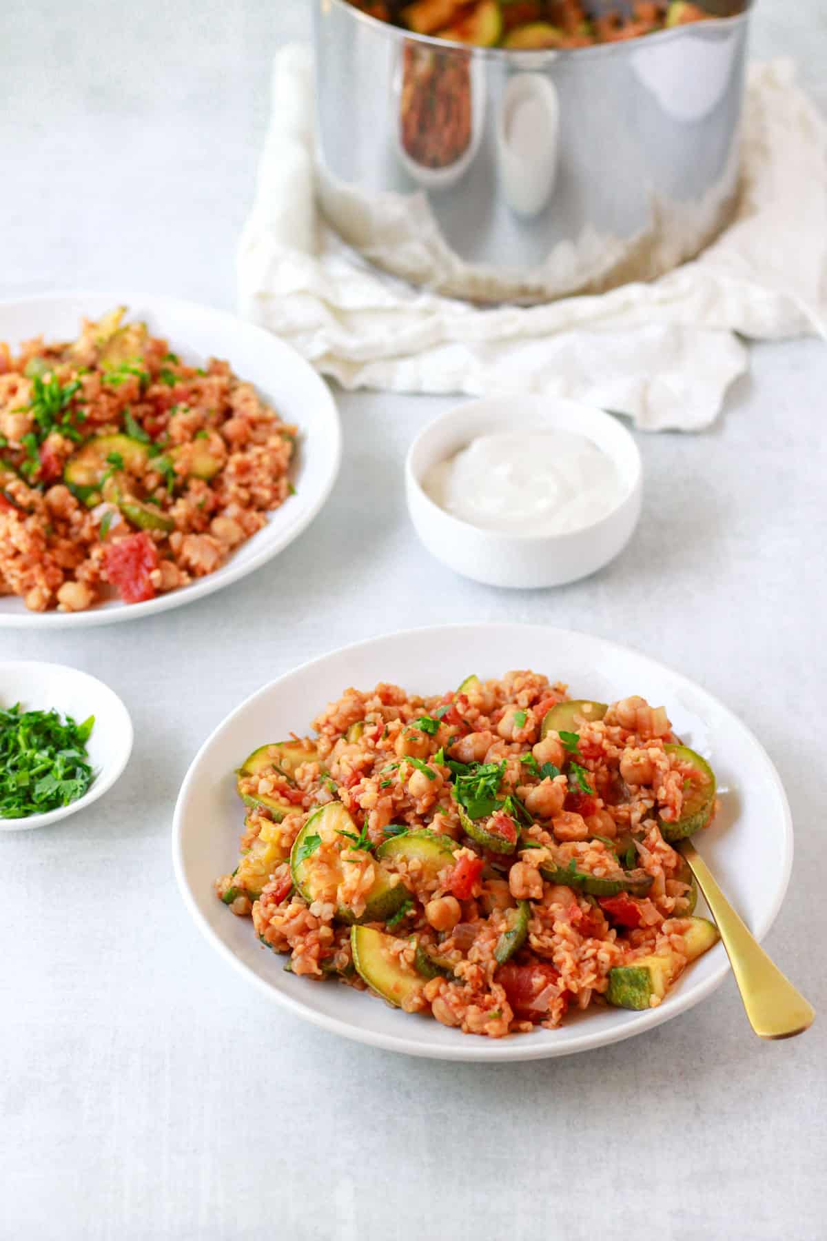 Two plates of tomato bulgur pilaf, shot at a 45 degree, with the pot in the background and yogurt for serving.
