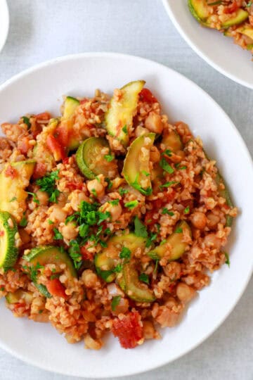 Image of bulgur pilaf with chickpeas, tomatoes, and squash, plated, topped with parsley, and cropped square.