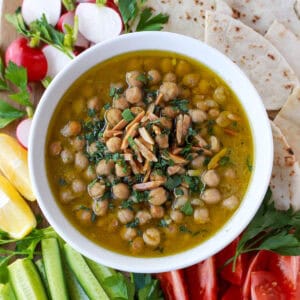 Balila, consisting of brothy, Lebanese chickpeas, surrounded by fresh crudités and pita bread for serving.