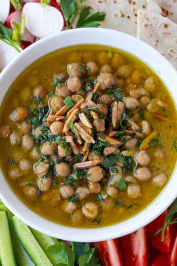 Balila, consisting of brothy, Lebanese chickpeas, surrounded by fresh crudités and pita bread for serving.