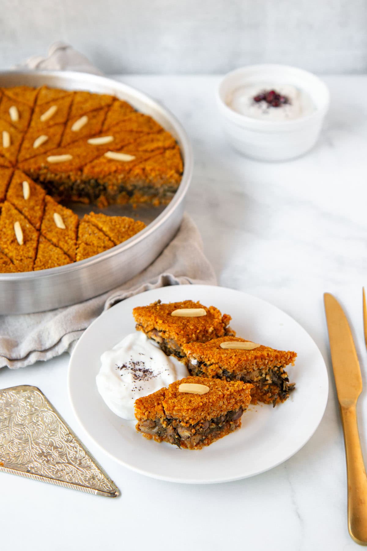 Diamond-shaped slices of pumpkin kibbeh served with yogurt and showing the rest of the baked kibbeh in the background.